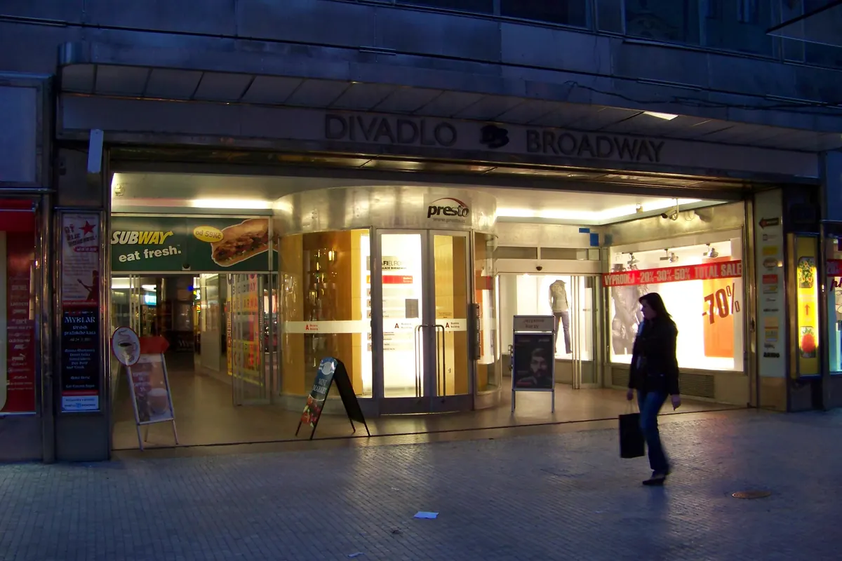The glass-fronted entrance of the Broadway arcade at dusk, with lit shop windows and a sign above reading Divadlo Broadway