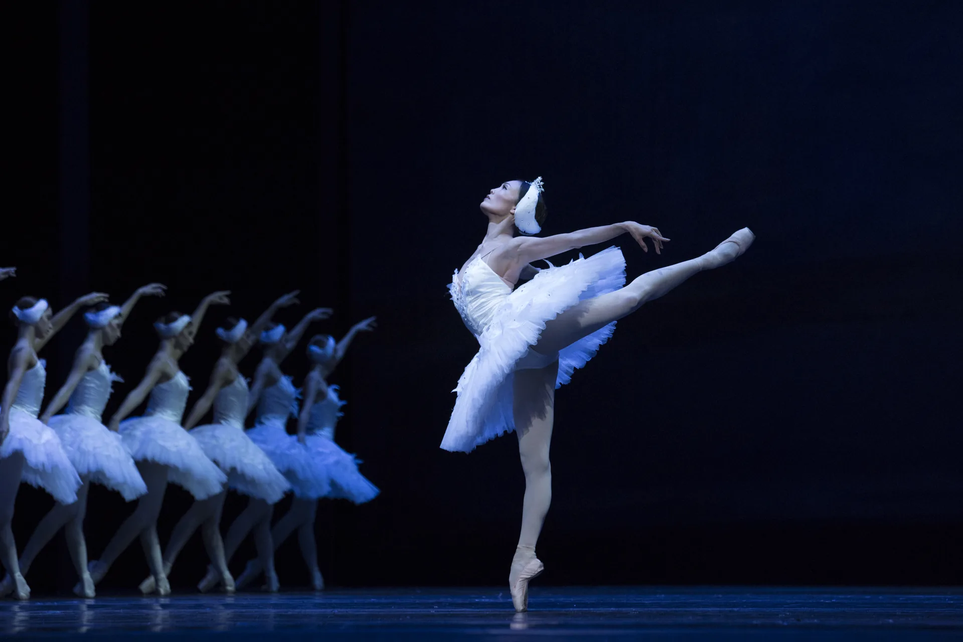 A ballerina in a white tutu and swan headdress balances on one pointe in a deep arabesque on a dark blue stage, with a line of swan corps dancers behind her, in a Swan Lake by another company
